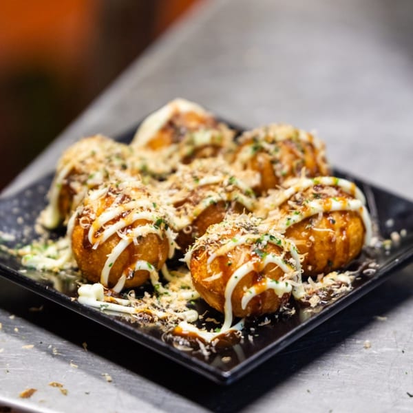 A black rectangular plate holds six golden-brown takoyaki, drizzled with white and brown sauces, and topped with green herbs.