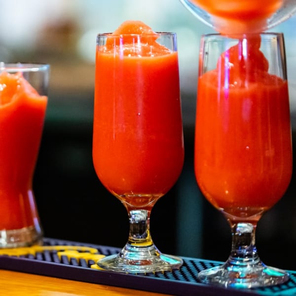 Three glasses of vibrant red slushy drinks, one being poured, on a dark bar counter.